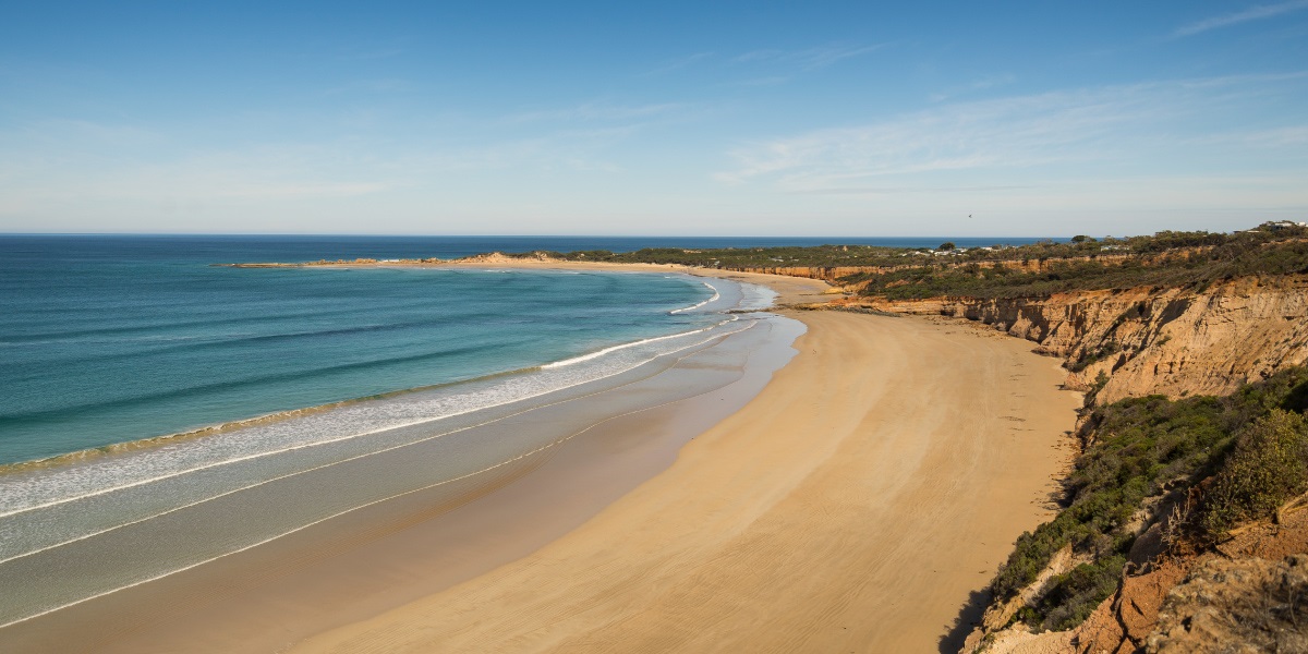 Landscape view of the Anglesea coastline with cliffs to the right, a stretch of sand in the centre and the blue ocean on the left.