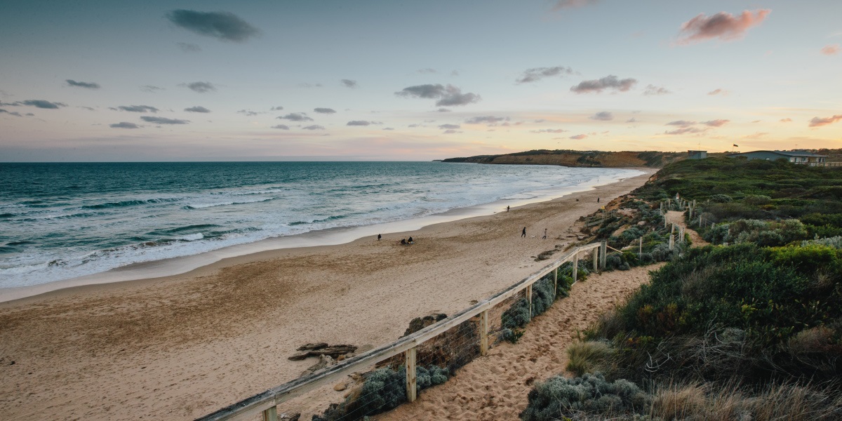 The view from the Jan Juc boardwalk looking left towards Bells Beach. The sun is setting to the right while the waves roll in to the sand.