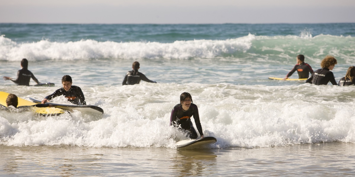A group of children in black wetsuits, surfing on foam boards in the waves close to the shore.