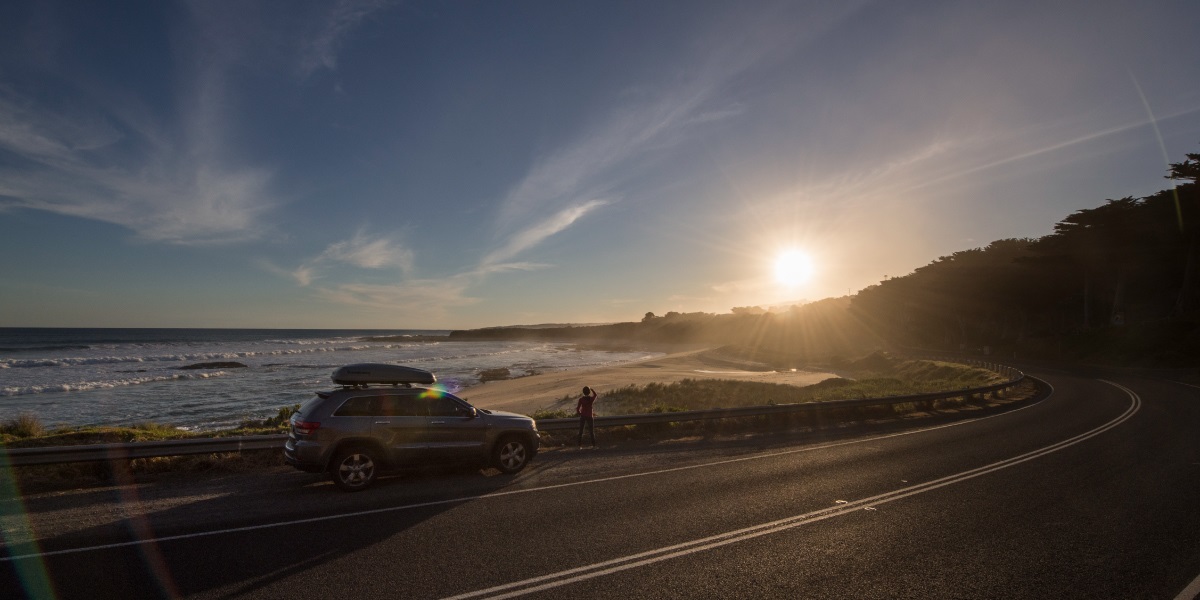 A car driving along the Great Ocean Road where the road meets the cliff, the sun is setting in the background.