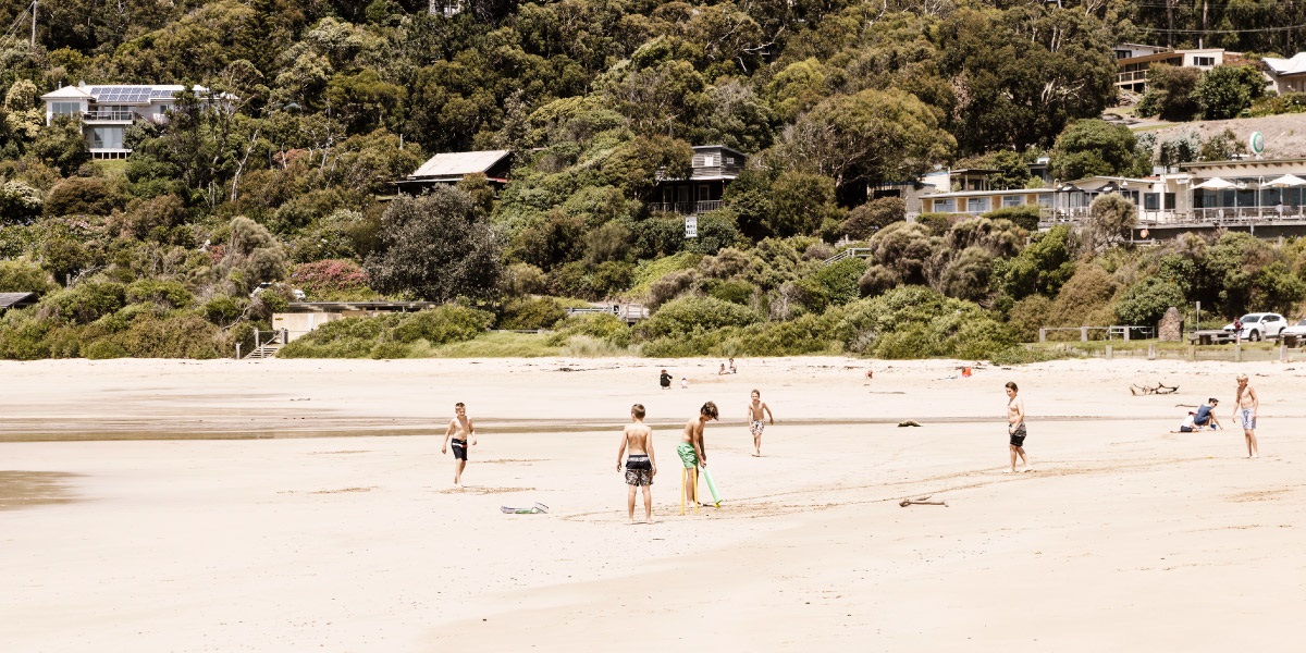 A group of people enjoying the summer sun on Wye River beach including a group of kids playing beach cricket. Trees cover the hills dotted with houses in the background.
