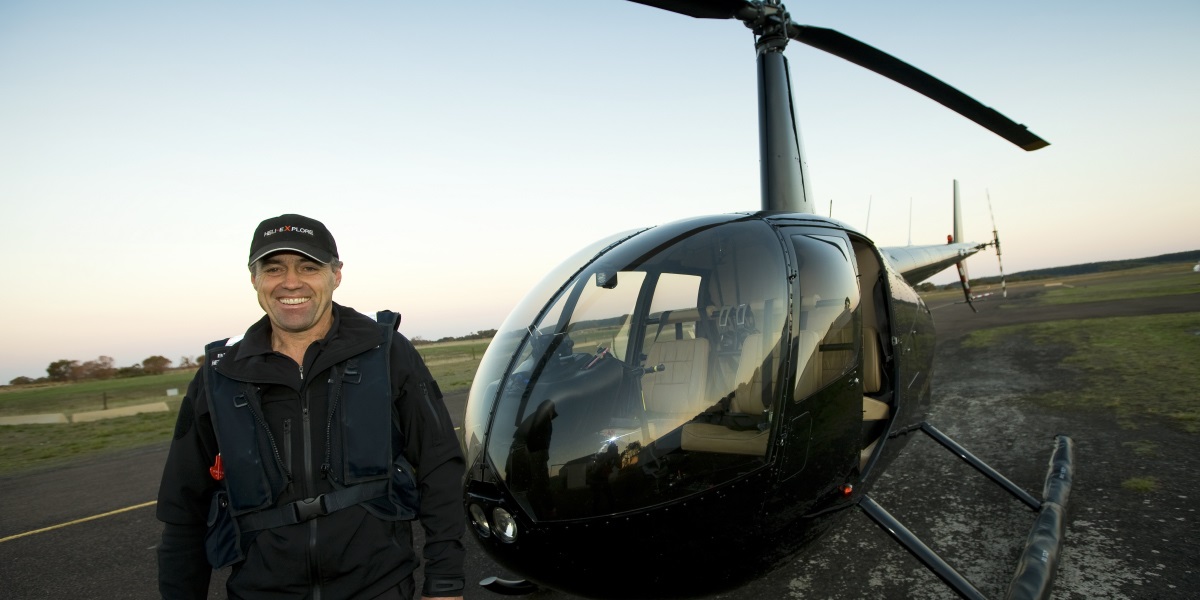A man dressed in a black flight suit stands smiling in front of a helicopter. A landing strip is in the background.