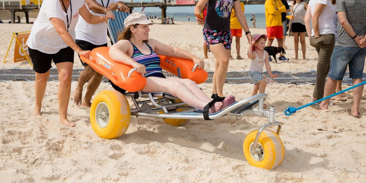 A person with a disability being pushed along the sand in a beach wheelchair. Everyone is smiling.