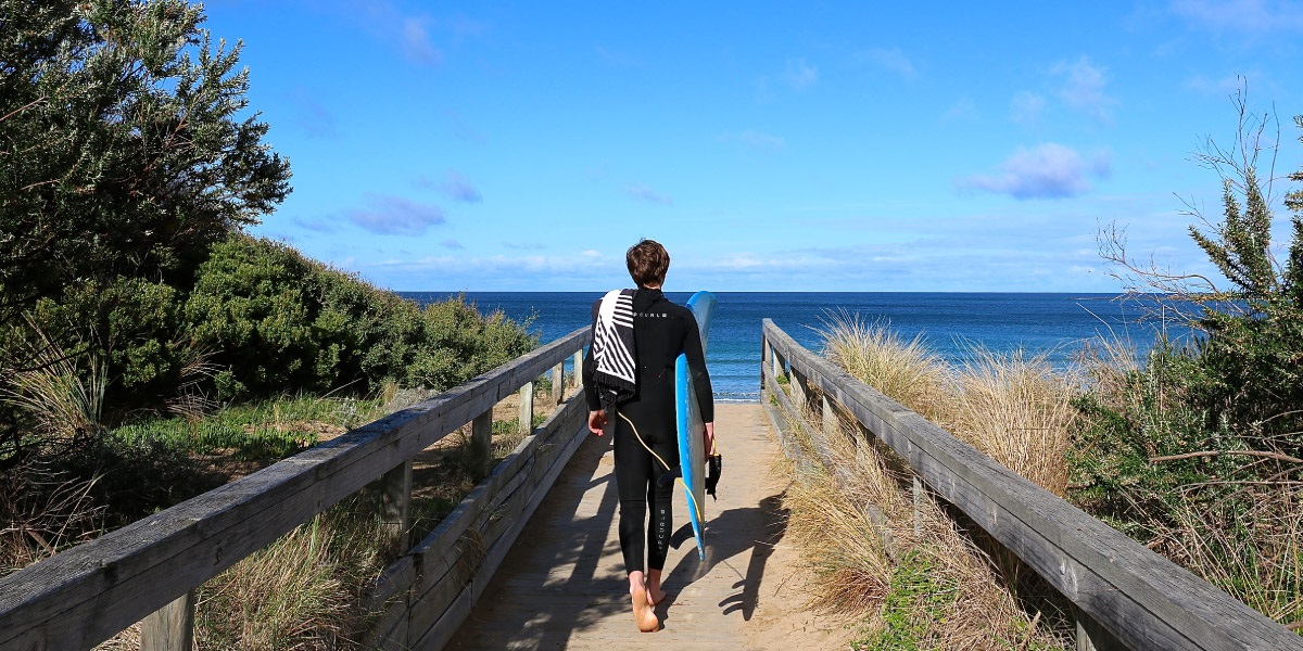 A person holding a foam surfboard walks down the boardwalk to the beach.