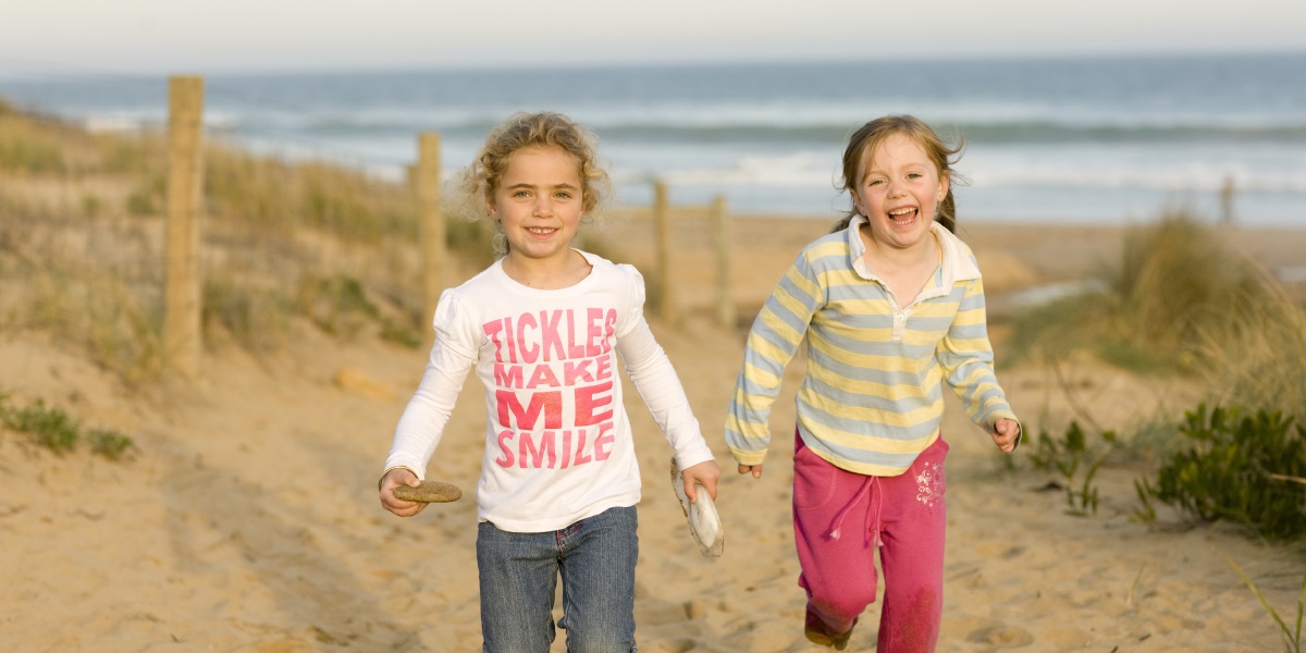 Two female children running up a sandy beach track holding hands. The ocean is blurred in the background.
