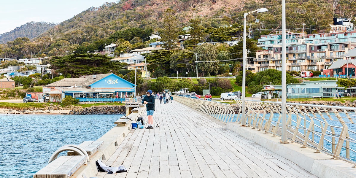 People fishing on Lorne pier with the Fisherman's Co-op and hills in the background.