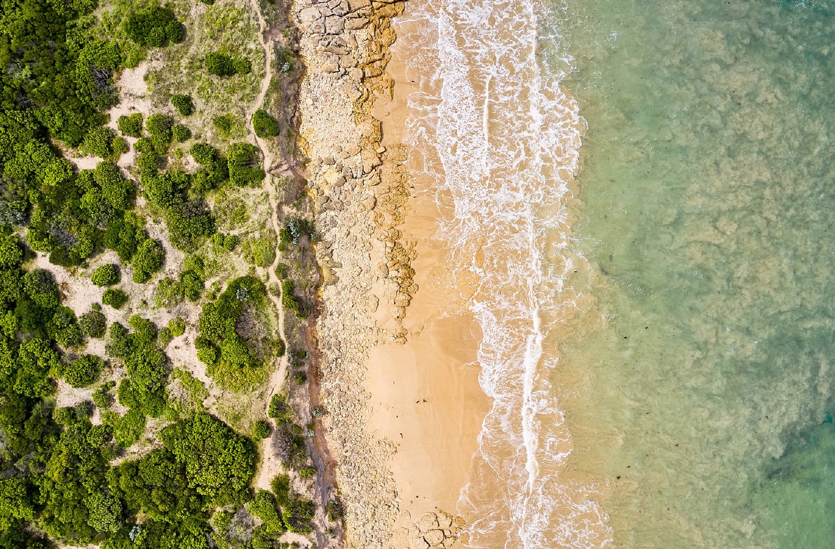 An aerial view of the light blue ocean with white tipped waves breaking on the sand. The beach is flanked by cliffs and green shrubs.