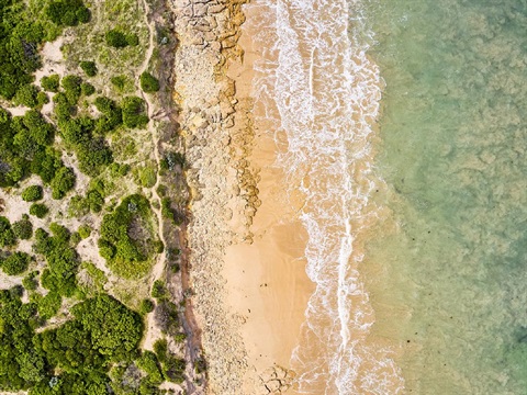 An aerial view of the light blue ocean with white tipped waves breaking on the sand.  The beach is flanked by cliffs and green shrubs.