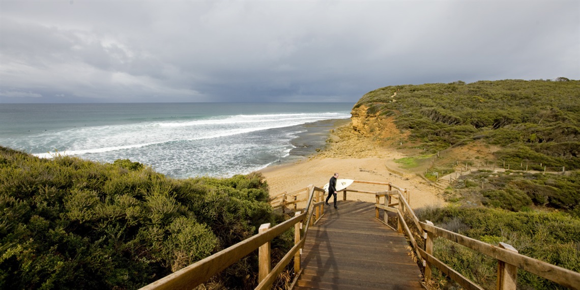 A surfer walking along the Aireys Inlet boardwalk down the stairs to the beach.  The sky is grey and the swell has many white peaks.